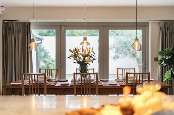 A dining area with a wooden table set for four, surrounded by chairs. There are hanging light fixtures above the table, and a vase with flowers in the center.