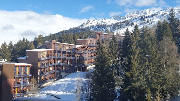 Et hotel med flere etager og balkoner, omgivet af sneklædte bjerge og fyrretræer.