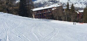 Une piste de ski enneigée, avec un bâtiment doté de nombreux balcons et un télésiège en arrière-plan.
