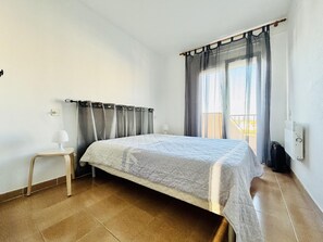 Ein Schlafzimmer mit Bett, Nachttisch, einem Fenster mit Vorhängen und Blick ins Freie.