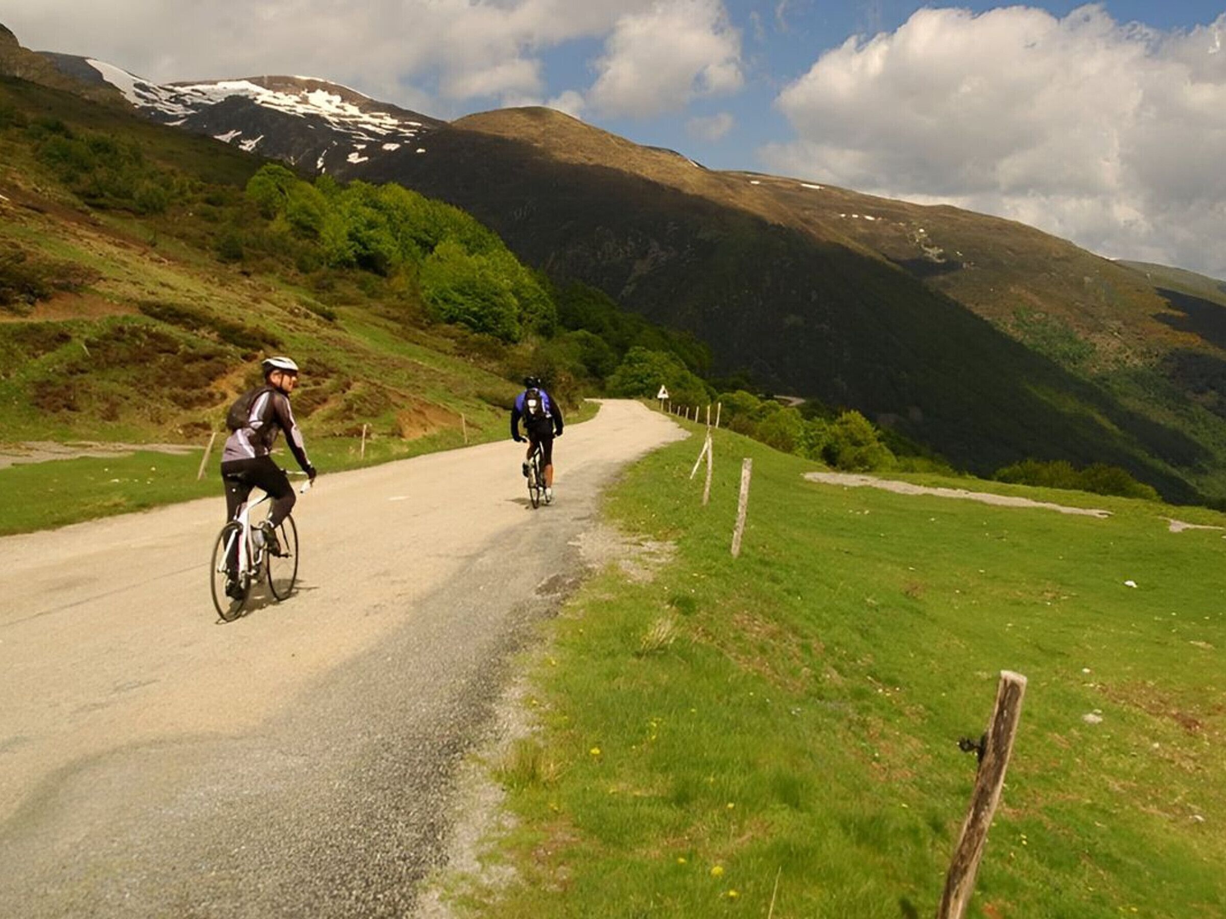 A gravel road with cyclists, surrounded by green hills and a mountain with snow patches.