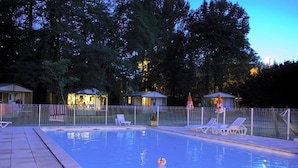 Piscina con sillones de descanso y sombrillas, rodeada por una valla y árboles.