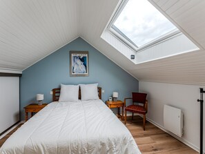 Ein Schlafzimmer mit schrägen Wänden, einem Dachfenster, einem Bett mit weißer Bettwäsche, zwei Nachttischen mit Lampen, einem Sessel und einem Heizkörper.