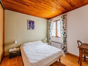 A bedroom with a wooden ceiling, a bed with a white quilt, a nightstand with a lamp, a window with patterned curtains, and a wooden chair. - 3-Bed House 100m from Beach (Biscarrosse)