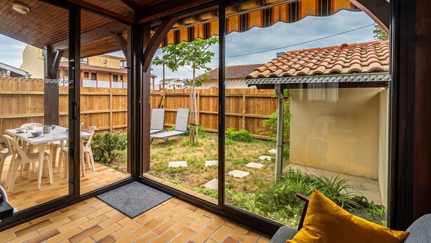 A covered patio with a brick floor, a table with chairs, a sofa with a cushion, and a view of a backyard with a wooden fence and a tiled roof structure. - 3-Bed House 100m from Beach (Biscarrosse)