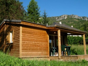 A wooden cabin with a porch and a table, surrounded by greenery and mountains in the background.