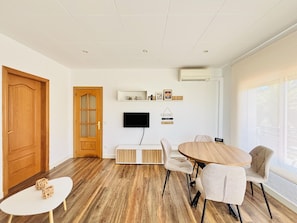 Ein modernes Wohnzimmer mit Holzboden, einem runden Esstisch mit Stühlen, einem weißen Wand-Fernseher und einer Holztür.