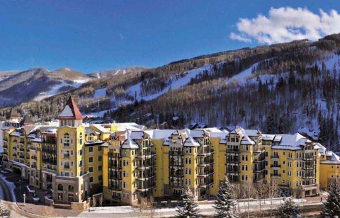 Ein mehrstöckiges Hotel mit gelber Fassade in einer Berglandschaft mit schneebedeckten Hängen und Kiefern.