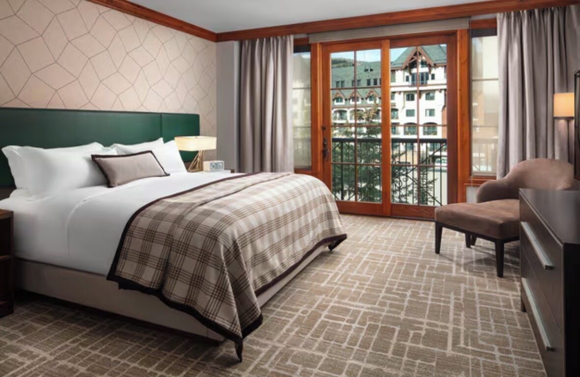 A hotel room with a large bed, a chair, a desk, and a view of a building through a glass door.