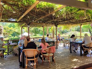 An outdoor patio area with wooden tables and wicker chairs, surrounded by lush greenery and a wooden pergola overhead. - Cozy Chalet with Terrace - Sleeps 5 (Pamiers)
