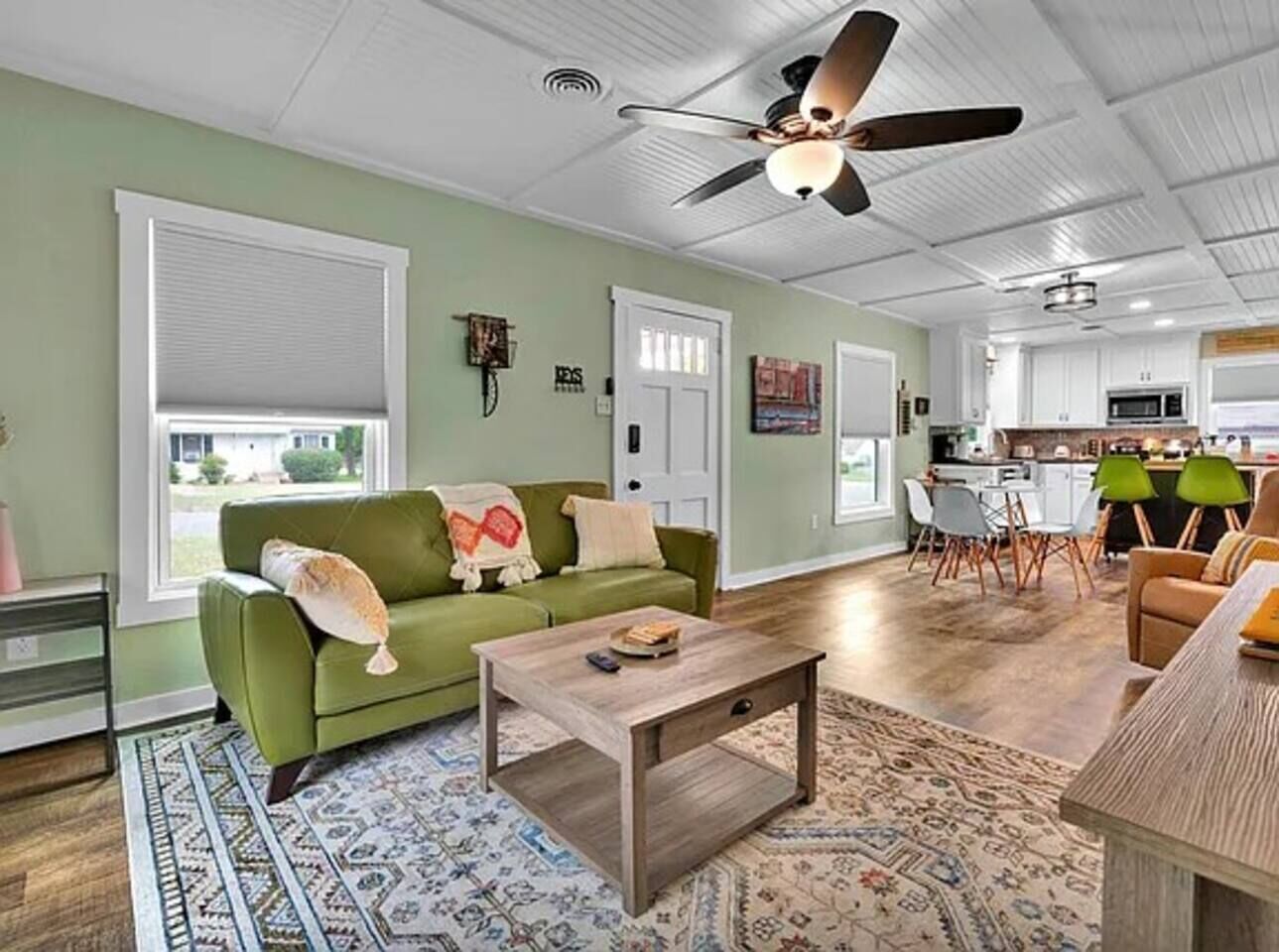 A living room with a green sofa, a coffee table, and a ceiling fan. There is a kitchen in the background with white cabinets and a dining area.