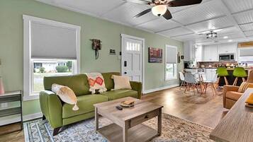 A living room with a green sofa, a coffee table, and a ceiling fan. There is a kitchen in the background with white cabinets and a dining area.