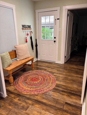 A wooden bench with cushions, a colorful woven rug, a white door with a window, and a 'WELCOME' sign.