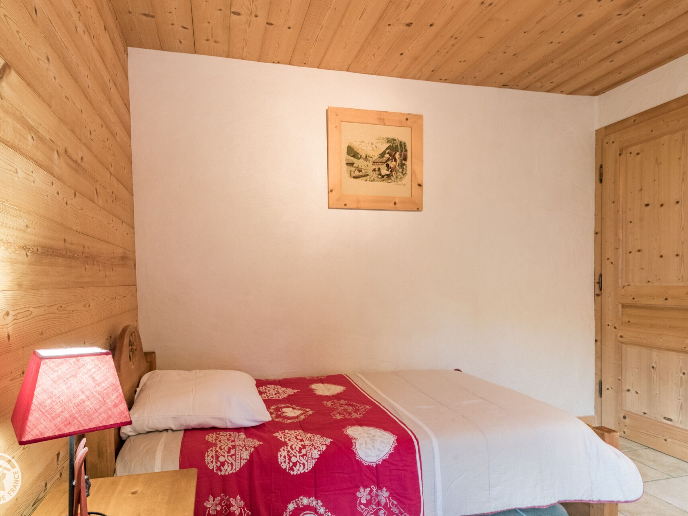 A bedroom with a wooden ceiling, a bed with a red and white bedspread, a white pillow, a wooden nightstand with a red lamp, and a framed picture on the wall.