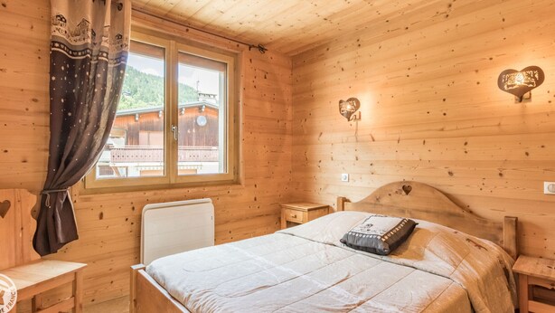 A wooden cabin bedroom with a bed, bedside tables, a window with curtains, and a view of a building outside. - Cozy apartment in Les Gets with parking - sleeps 5 (Les Gets)