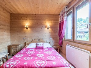 A wooden cabin bedroom with a bed, two bedside tables, a nightstand, a window with curtains, and a view of a forested area. - Cozy apartment in Les Gets with parking - sleeps 5 (Les Gets)