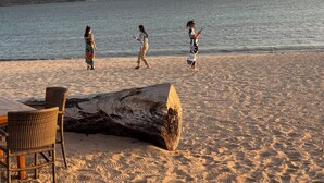 Een zandstrand met een grote boomstam, rieten stoelen en een tafel.