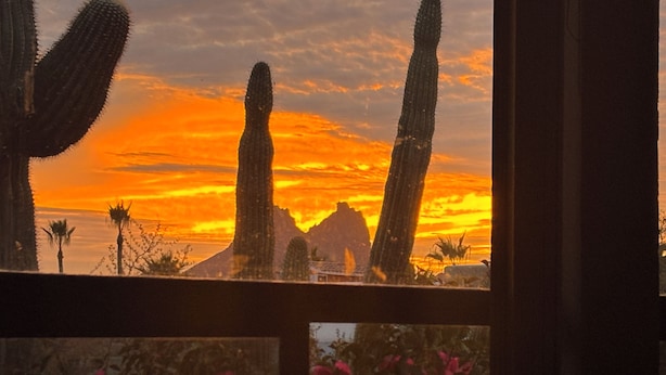 Een zonsondergang door een raam, met cactussen en een berg op de achtergrond.