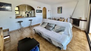 Ein Wohnzimmer mit Sofa, Couchtisch, Küchenzeile mit Theke und Barhocker sowie Kamin.