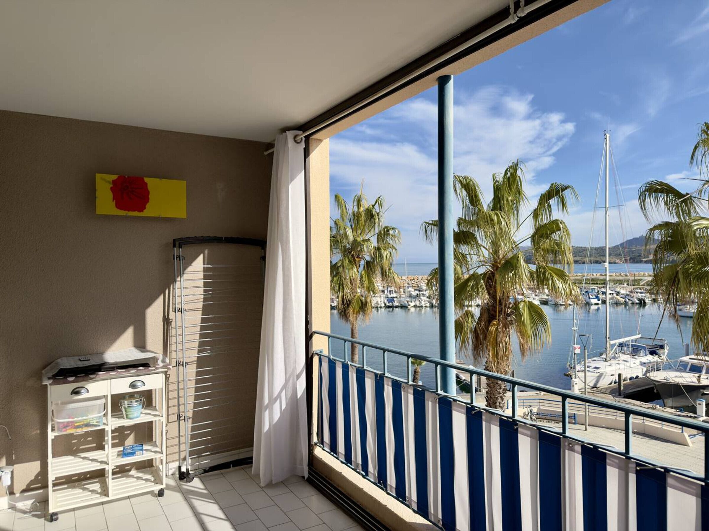 Een balkon met uitzicht op een jachthaven, een wit schap met spullen, en een geel bord aan de muur.