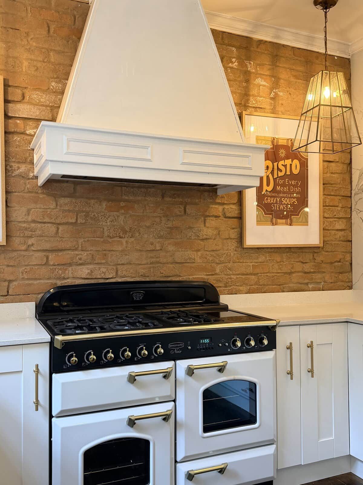 A kitchen with a white stove, oven, and cabinetry, a brick wall, and a hanging light fixture.