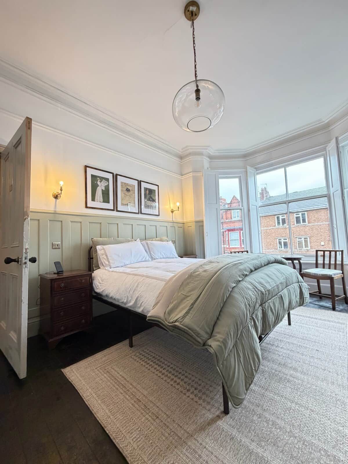 A bedroom with a bed, a dresser, a nightstand, a lamp, and a window with a view of buildings.