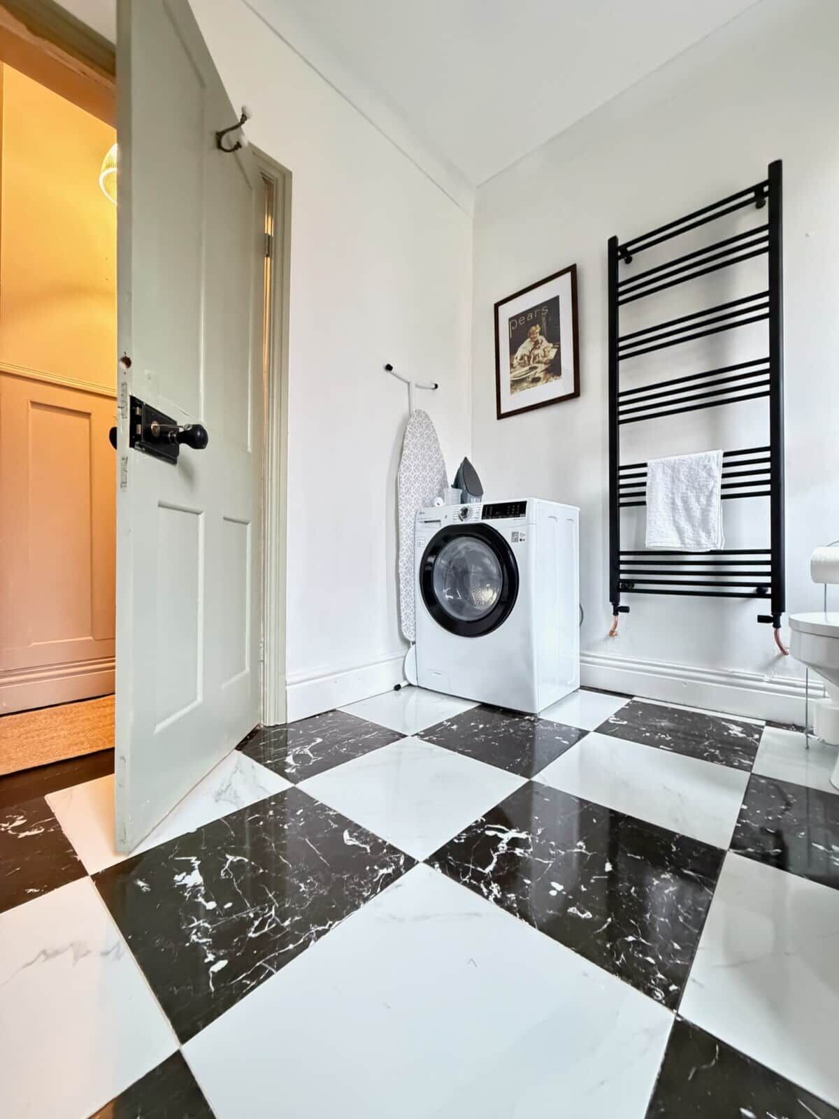 A laundry area with a washing machine, ironing board, and a black towel rack.
