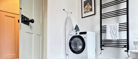 A laundry area with a washing machine, ironing board, and a black towel rack.
