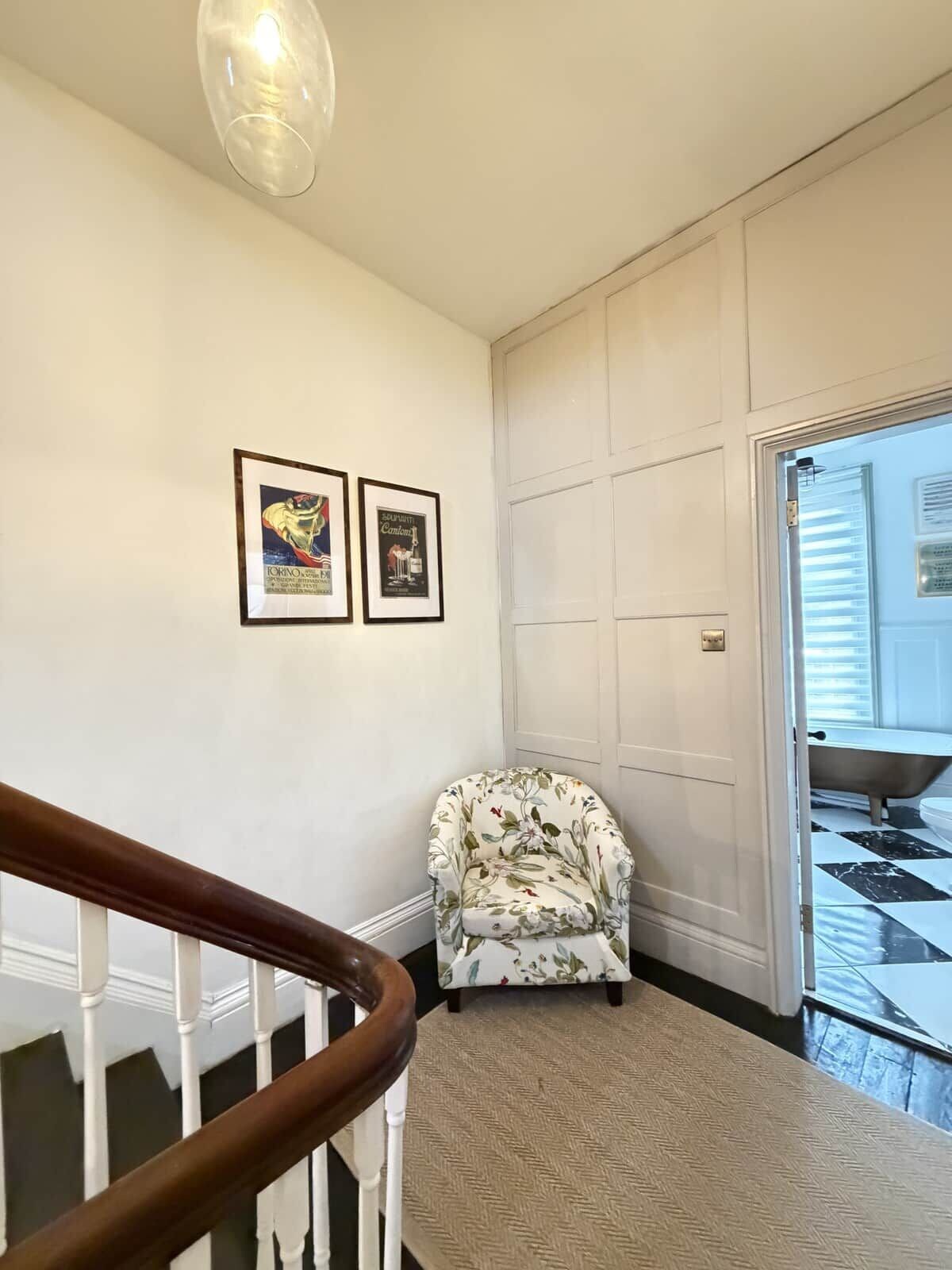 A staircase with a wooden handrail leading to a landing. On the landing, there is a floral-patterned chair and a framed picture on the wall.