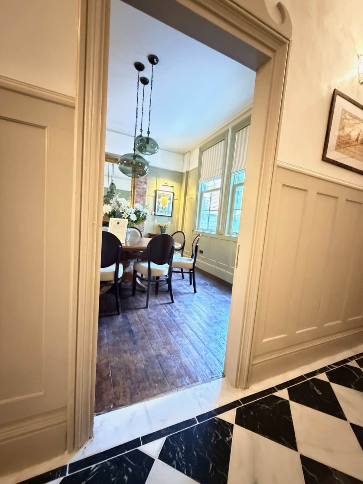 En gang der fører til et spiseområde med trægulv, en lysekrone og et indrammet billede på væggen.