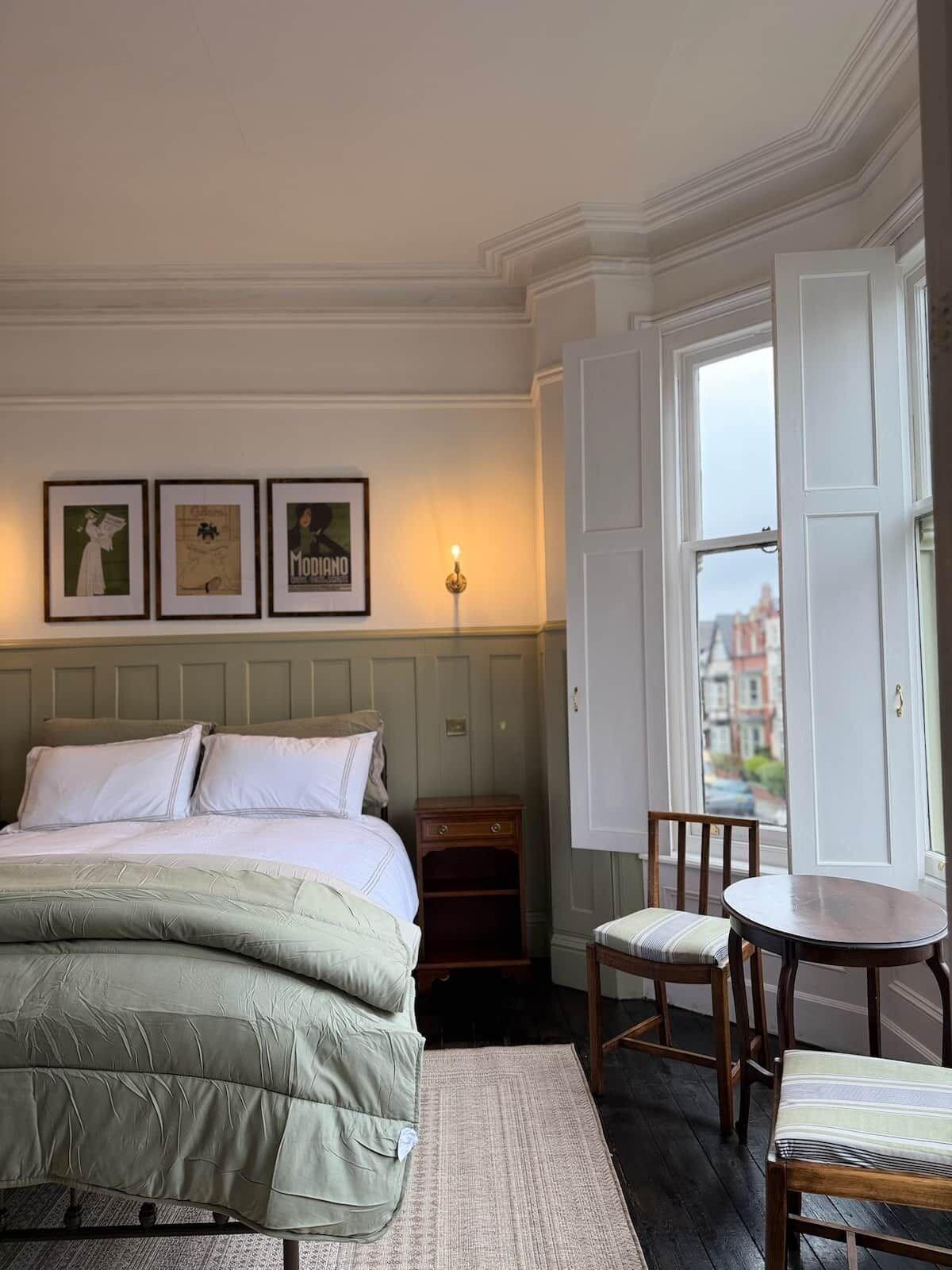 A hotel room with a bed, a small table, two chairs, a nightstand, and a window with a view of buildings.