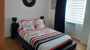 A neatly made bed with striped bedding, a round wall decoration, a small table with a lamp, and a window with blinds. - Family home close to the sea (Penmarch)