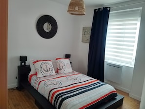 Ein ordentlich bezogenes Bett mit Streifenmuster, eine runde Wanddekoration, ein kleiner Tisch mit einer Lampe und ein Fenster mit Jalousien.