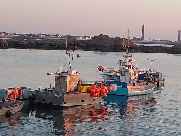Ein Hafen mit mehreren vertäuten Booten, ein Leuchtturm im Hintergrund und ruhiges Wasser, das die Boote spiegelt.