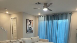 Una sala de estar con un sofá modular, un ventilador de techo, un ventanal con cortinas y un cuadro en la pared.