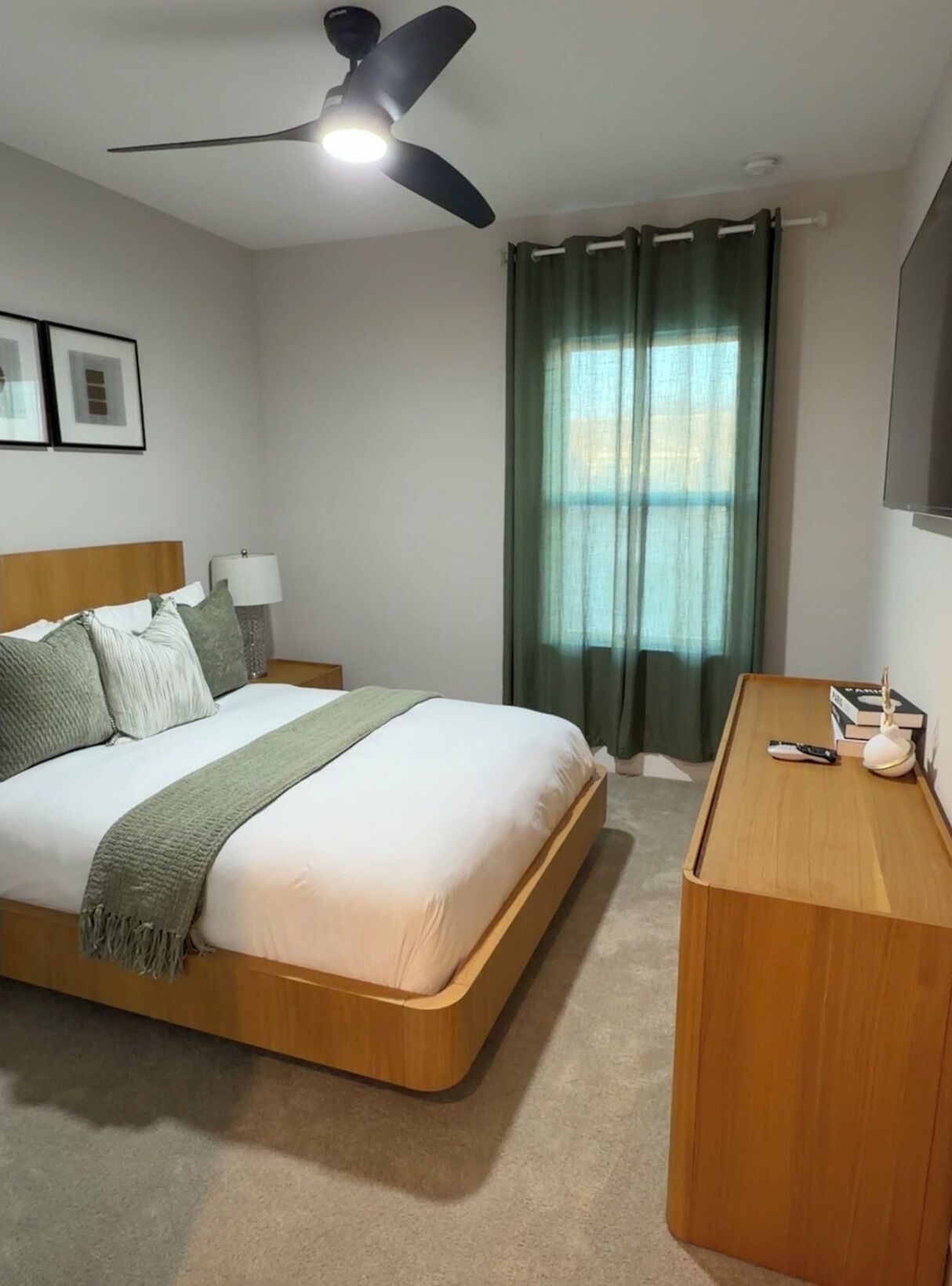 A hotel room with a bed, a desk, a ceiling fan, a window with curtains, and framed pictures on the wall.