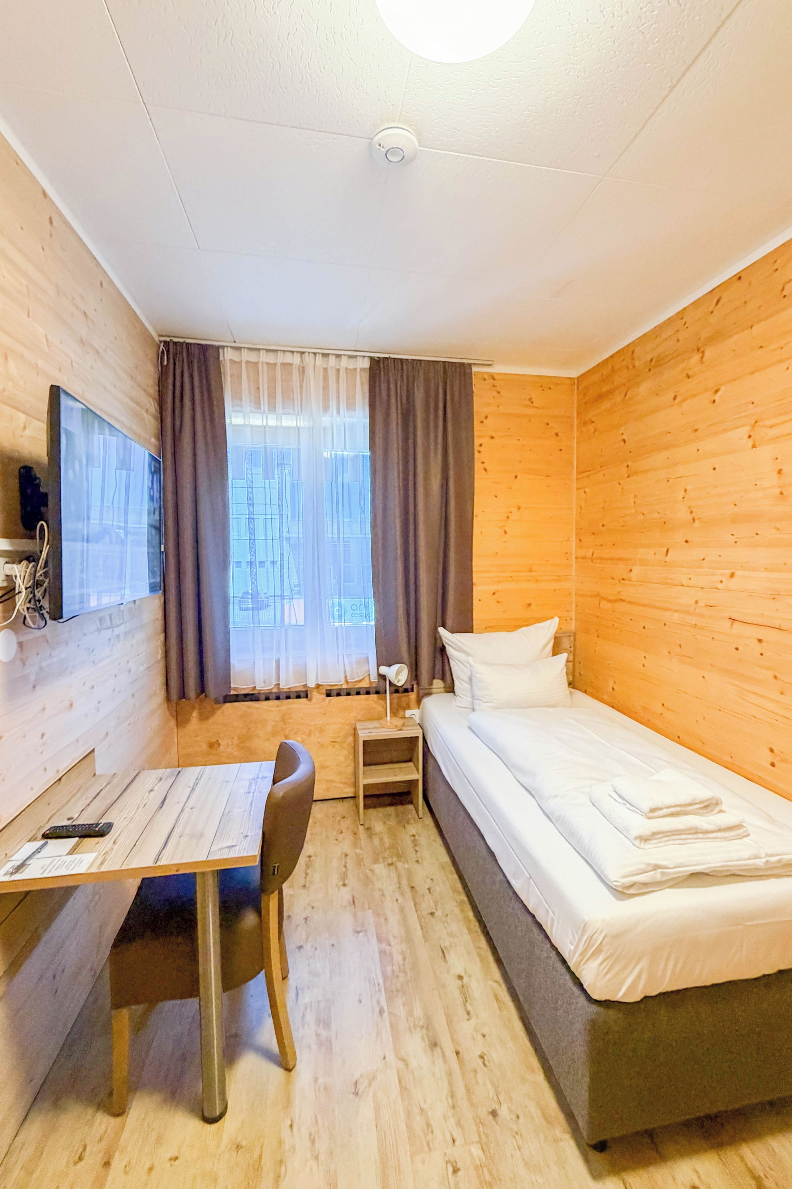 Ein Zimmer mit einem Bett, einem Schreibtisch, einem Stuhl, einem Fernseher und einem fensterbepflanzten Fenster.