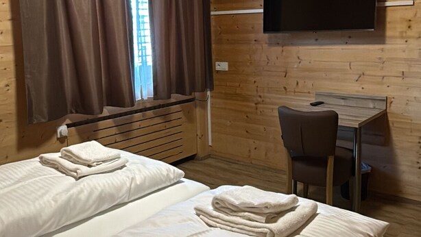 Ein Zimmer mit zwei Betten, einem Schreibtisch, einem Stuhl, einem an der Wand befestigten Flachbildfernseher und einem Fenster mit Vorhängen.