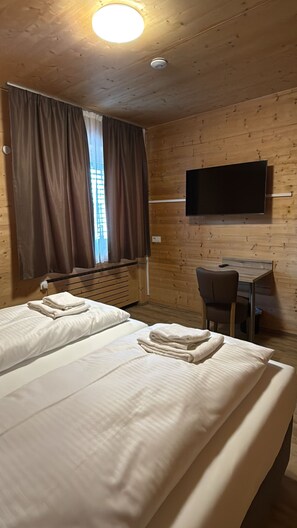 A room with two beds, a desk, a chair, a flat-screen TV mounted on the wall, and a window with curtains. - URBAN OAK Smart Stay (Cologne)