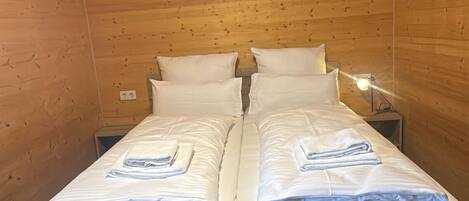 A double bed with white linens and folded towels, a bedside table with a lamp, and wooden paneling on the walls.