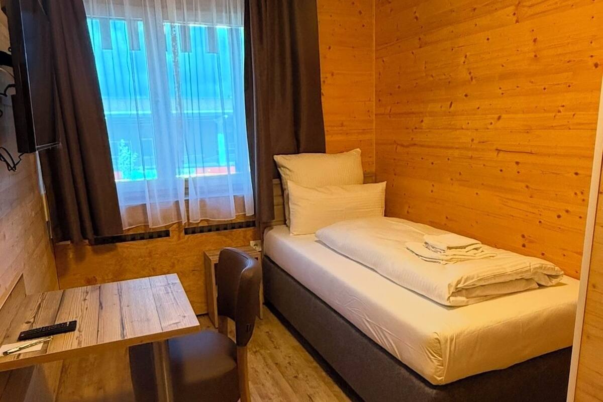 A small, cozy room with a wooden floor, a bed, a desk with a chair, and a window with curtains.