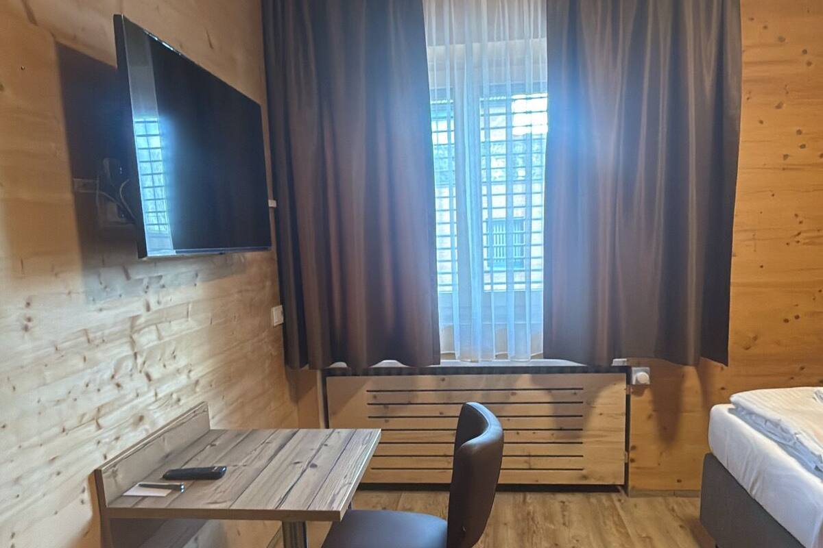 A wooden cabin room with a bed, a table with a chair, a TV mounted on the wall, and a window with curtains.