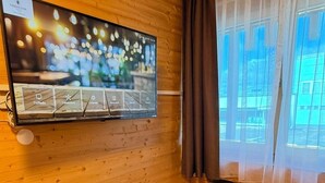 Ein Zimmer mit Holzwänden, an der Wand montiertem Fernseher, Schreibtisch, Stuhl und einem Fenster mit Vorhängen.