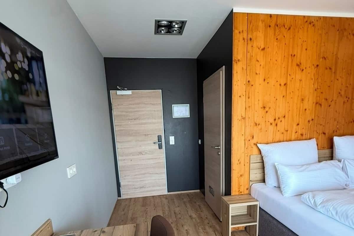 A modern hotel room with a wooden floor, a bed with white linens, a desk with a chair, a television, and a wardrobe.