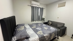 Ein Schlafzimmer mit einem Bett, einem grauen Sofa, einem Fenster mit Vorhängen, einer Klimaanlage und einem Wandventilator.