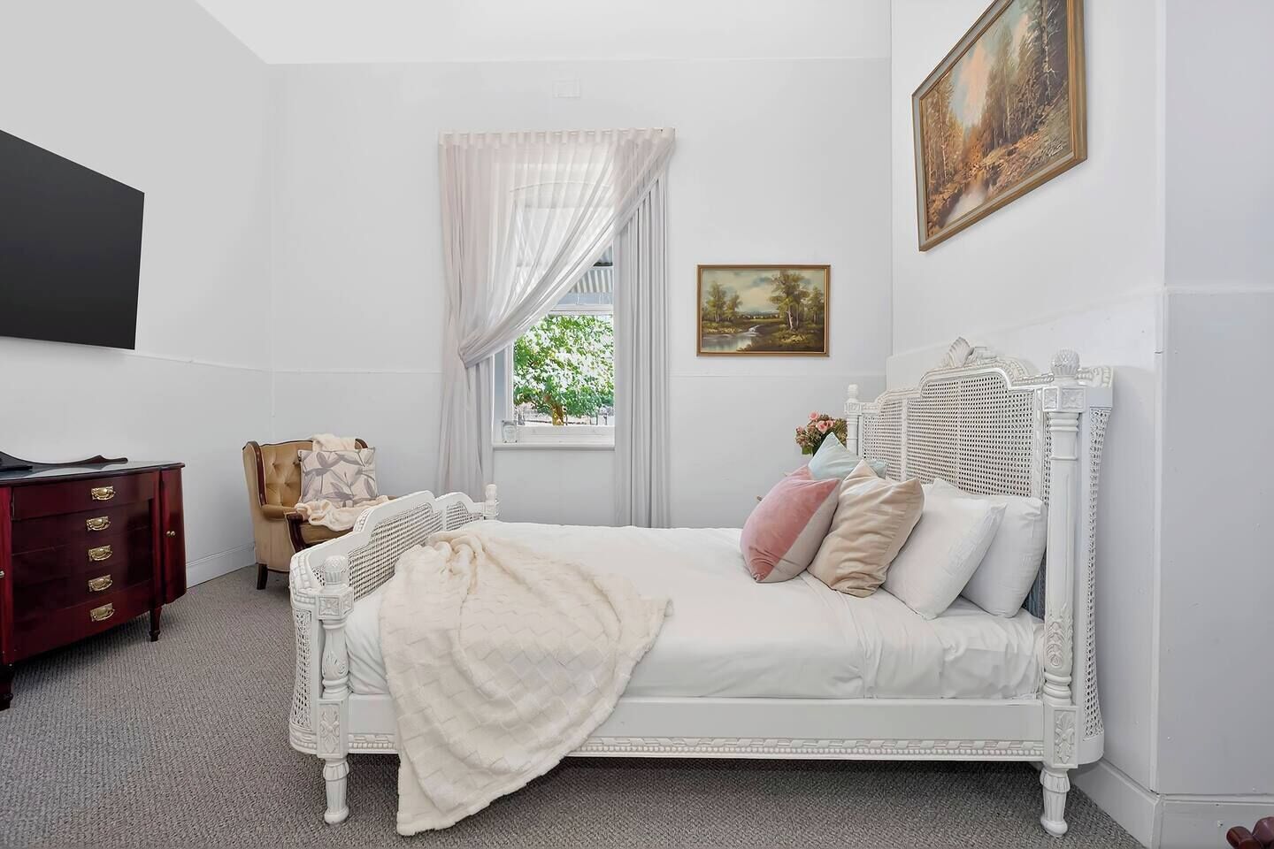 Une chambre à coucher décorée dans des tons de blanc, avec un lit, une commode, une chaise et une fenêtre avec des rideaux.