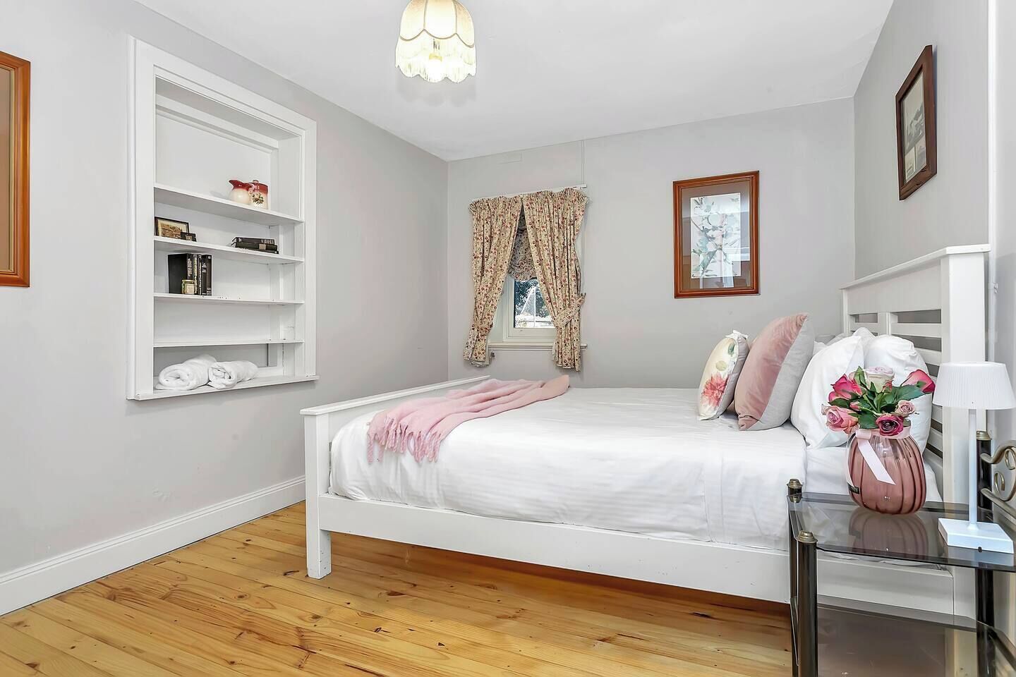 Une chambre avec un lit blanc, un plancher en bois, une armoire blanche et une fenêtre ornée de rideaux à motifs floraux.