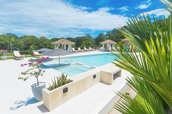 A swimming pool with lounge chairs and thatched umbrellas, surrounded by greenery and a clear sky.