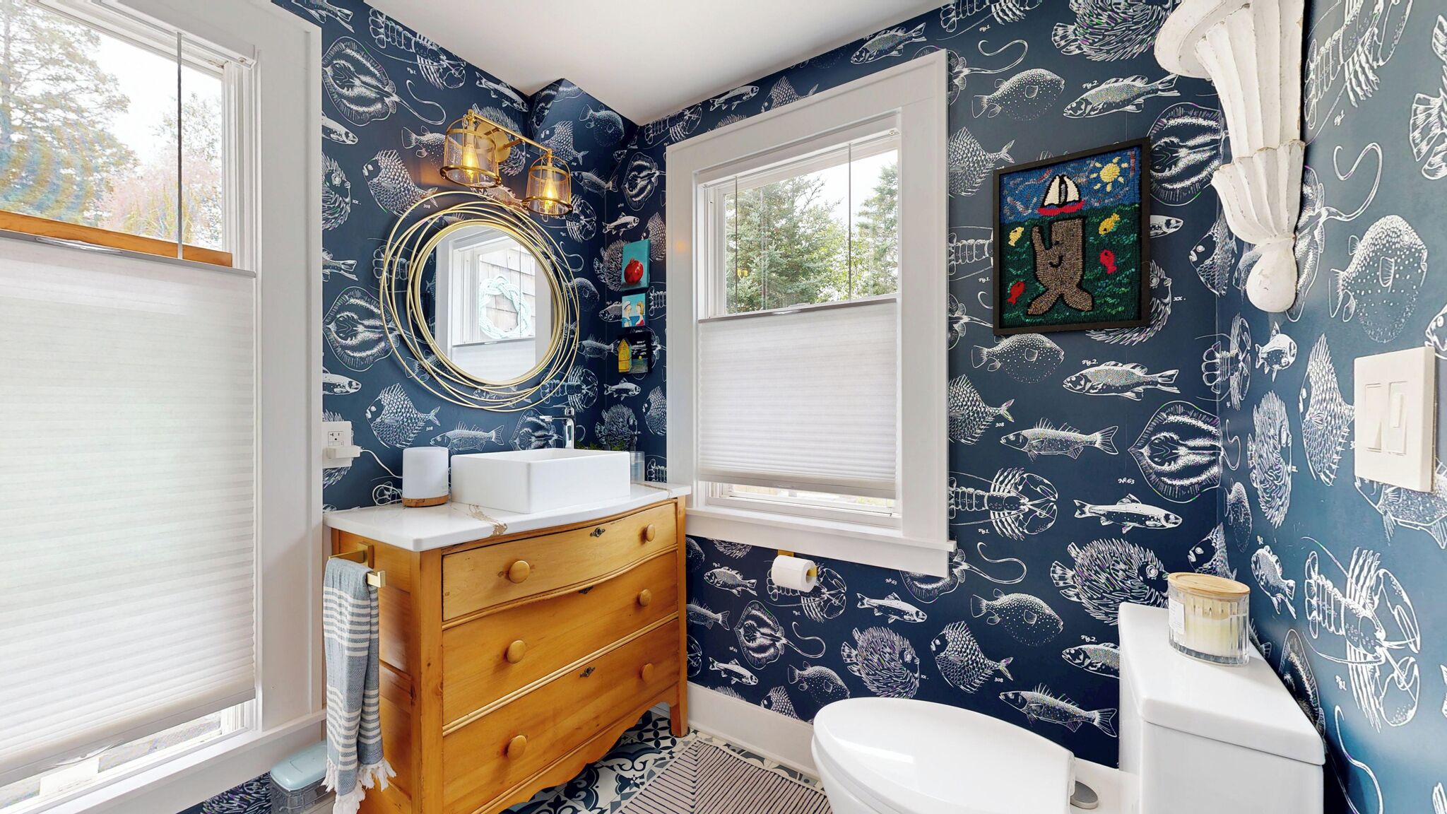 A bathroom with a nautical theme, featuring fish patterns on the walls, a wooden vanity, a round mirror, and a toilet.