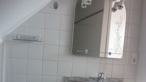 Un baño con lavamanos blanco, espejo y paredes con azulejos. Hay un perchero y una zona de ducha con puerta de cristal.